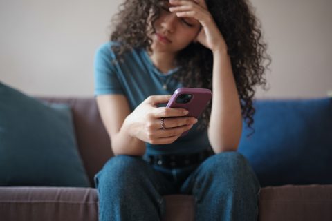 szomorú, fiatal hosszú hajú nő lila tokos mobiltelefonnal a kezében, a fejét fogja