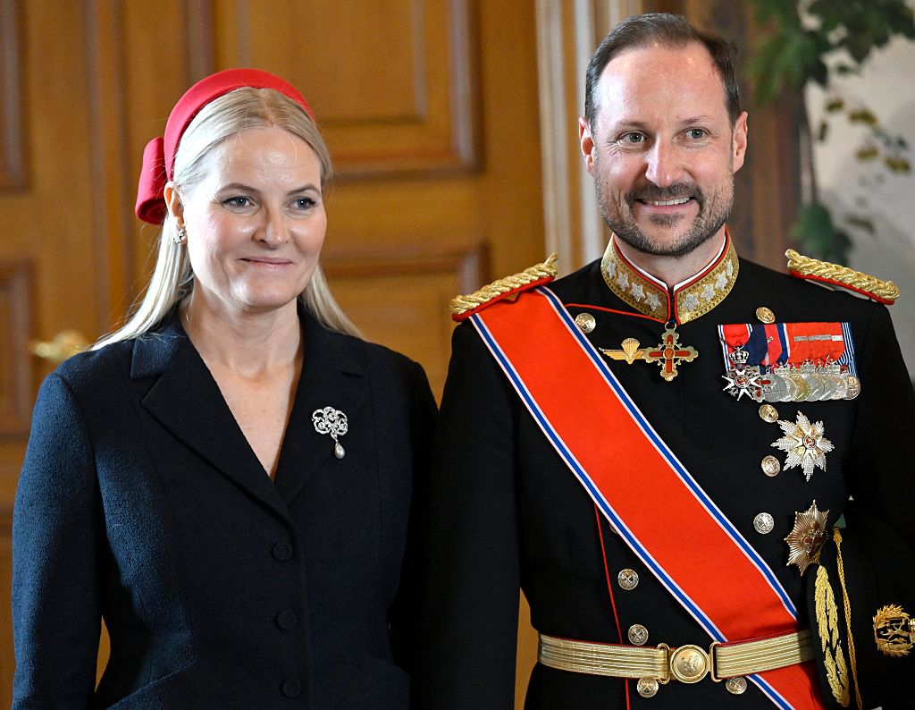 Haakon hivatalos eseményhez öltözve és Mette-Marit piros kalapban és fekete ruhában