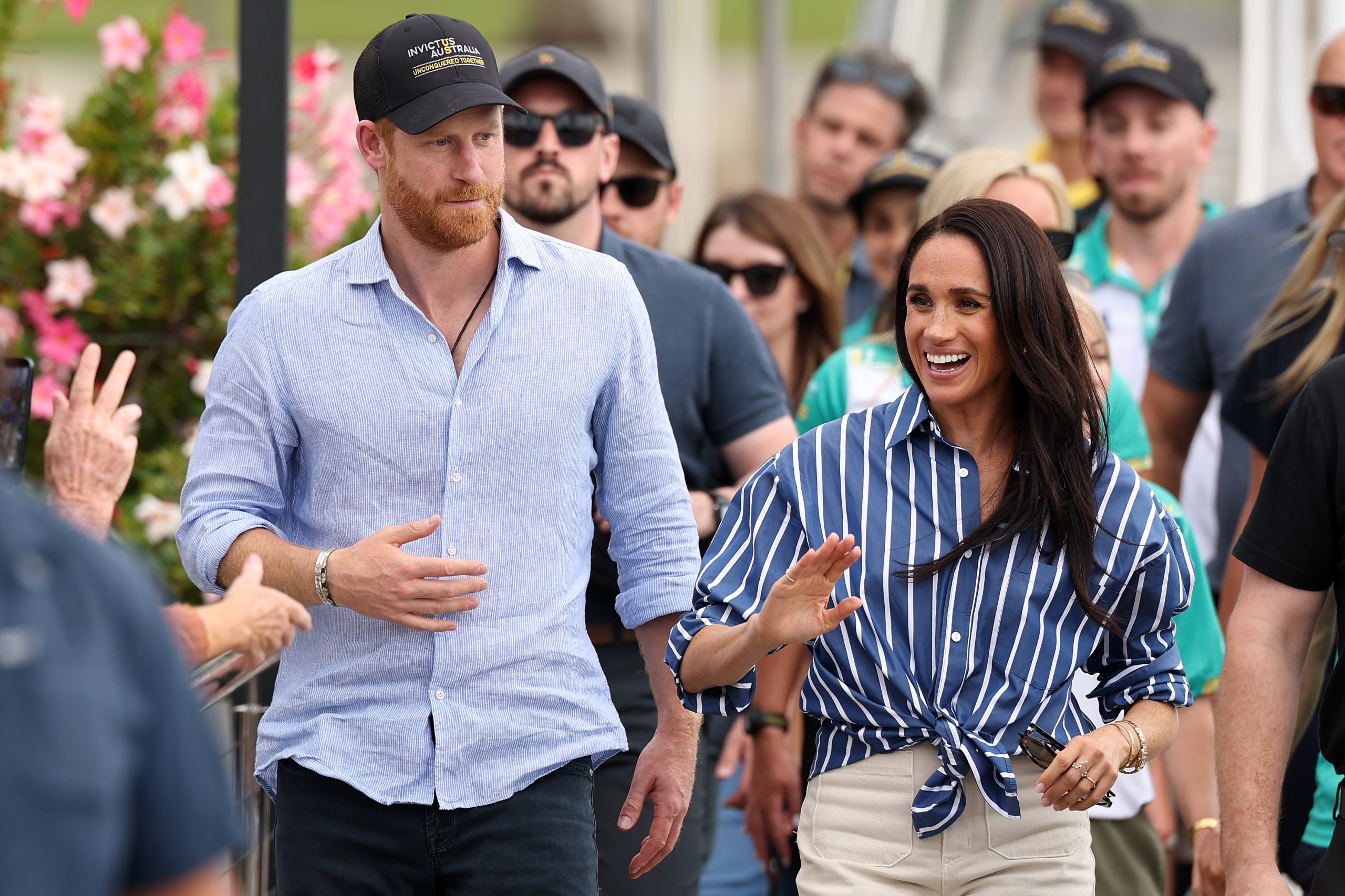 Harry herceg kék ingben és baseball sapkában, mellette Meghan kék fehér csíkos ingben, nevetve