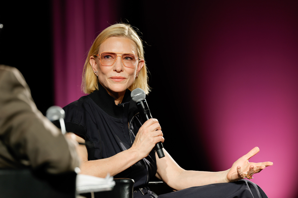 cannes-2026-cate-blanchett | Marie Claire Cate Blanchett sötétkék felsőben és narancssárga szemüvegben, mikrofonnal a kezében.