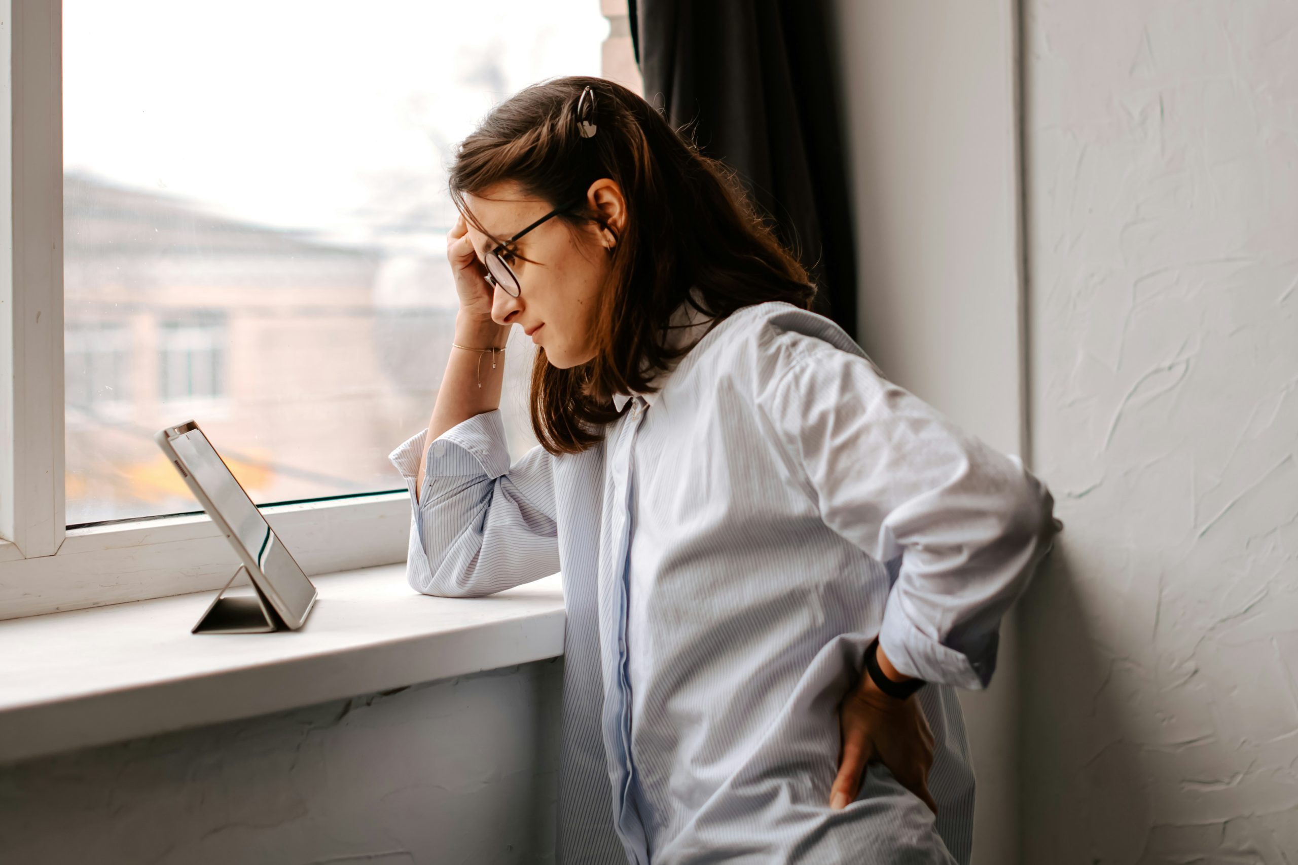 Stresszes fiatal nő fejét fogva áll az ablak előtt
