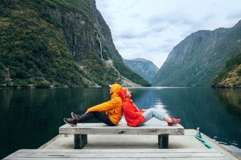 Egy férfi narancsszínű kabátban és egy nő piros kabátban egymás hátát támasztva ül Norvégiában egy fjord partján lévő padon
