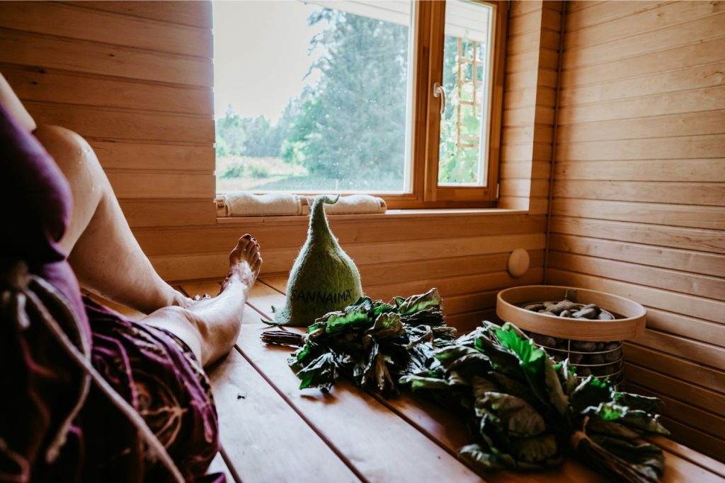norvegia-hosszu-elet-szauna | Marie Claire Nő szaunában ül, mellette gyógynövények, szaunasapka, kilátás ablakon az erdőre