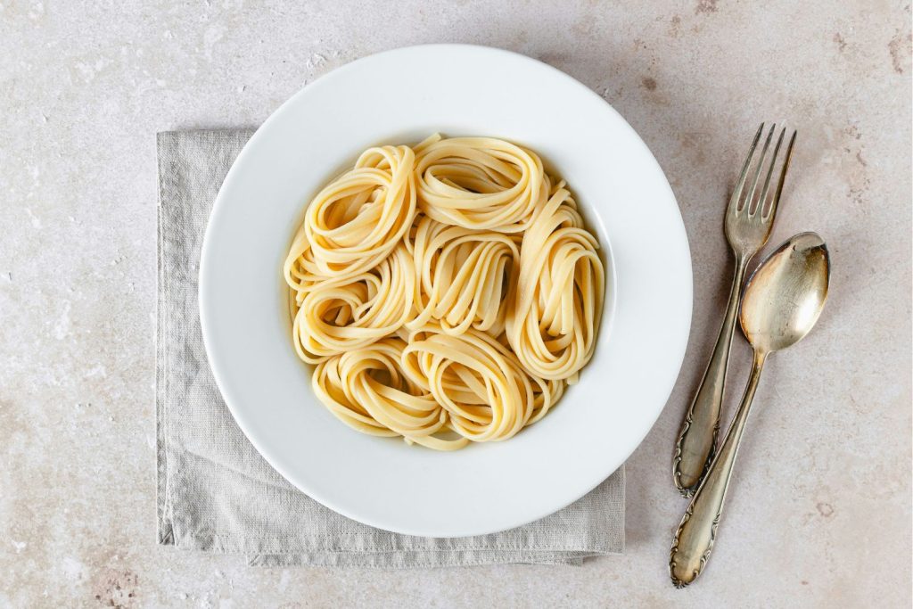 Fehér tányérban spagetti szürke asztalon, mellette rusztikus evőeszközök