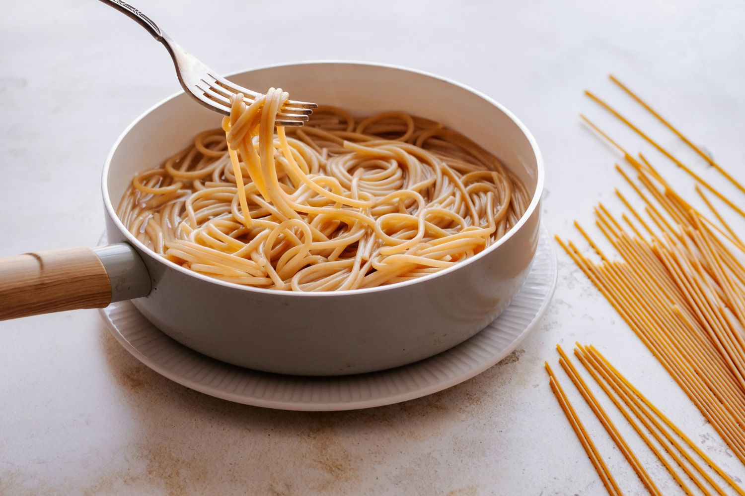 Spagetti serpenyőben, villával kivesz egy adagot, mellette az asztalon száraztészta