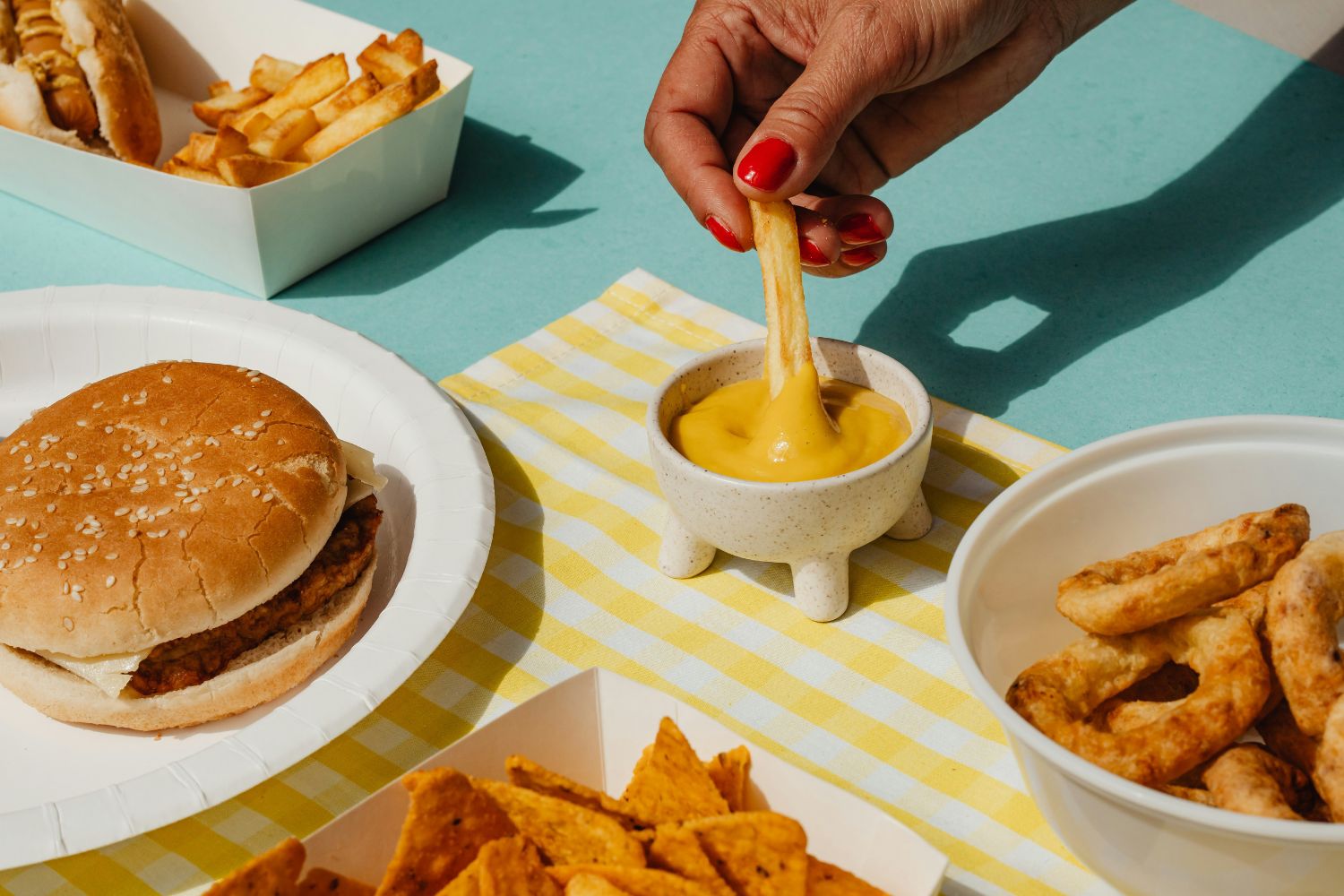 Sárga csíkos asztalterítőn junk food burgerrel, sült krumplit mustárba mártó pirosra lakkozott körmű női kéz, chips kék asztalon