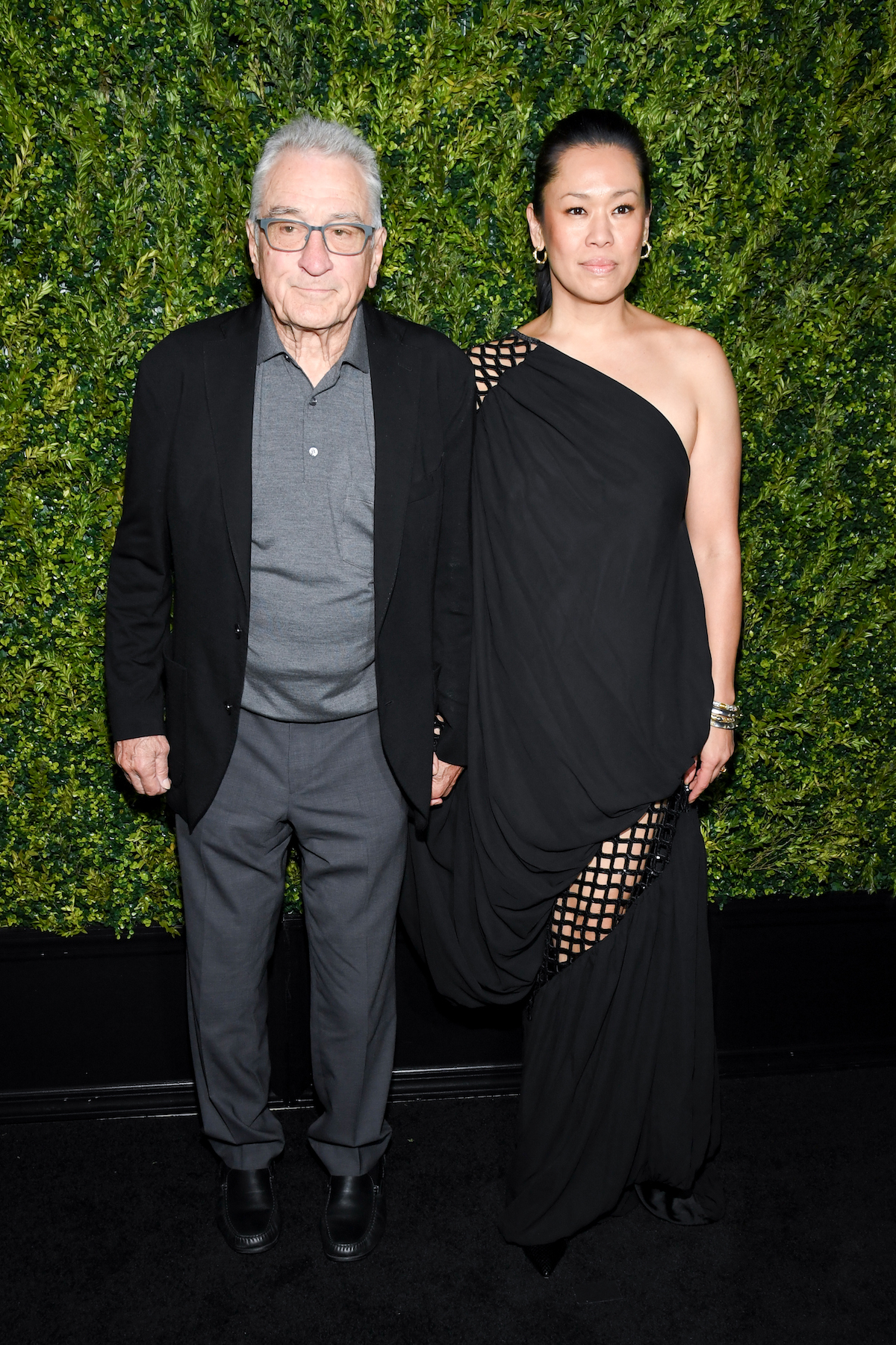 robert-de-niro-tiffany-chen | Marie Claire Robert De Niro zakóban és szürke ingben, és Tiffany Chen fekete félvállas ruhában, kézen fogva zöld háttér előtt