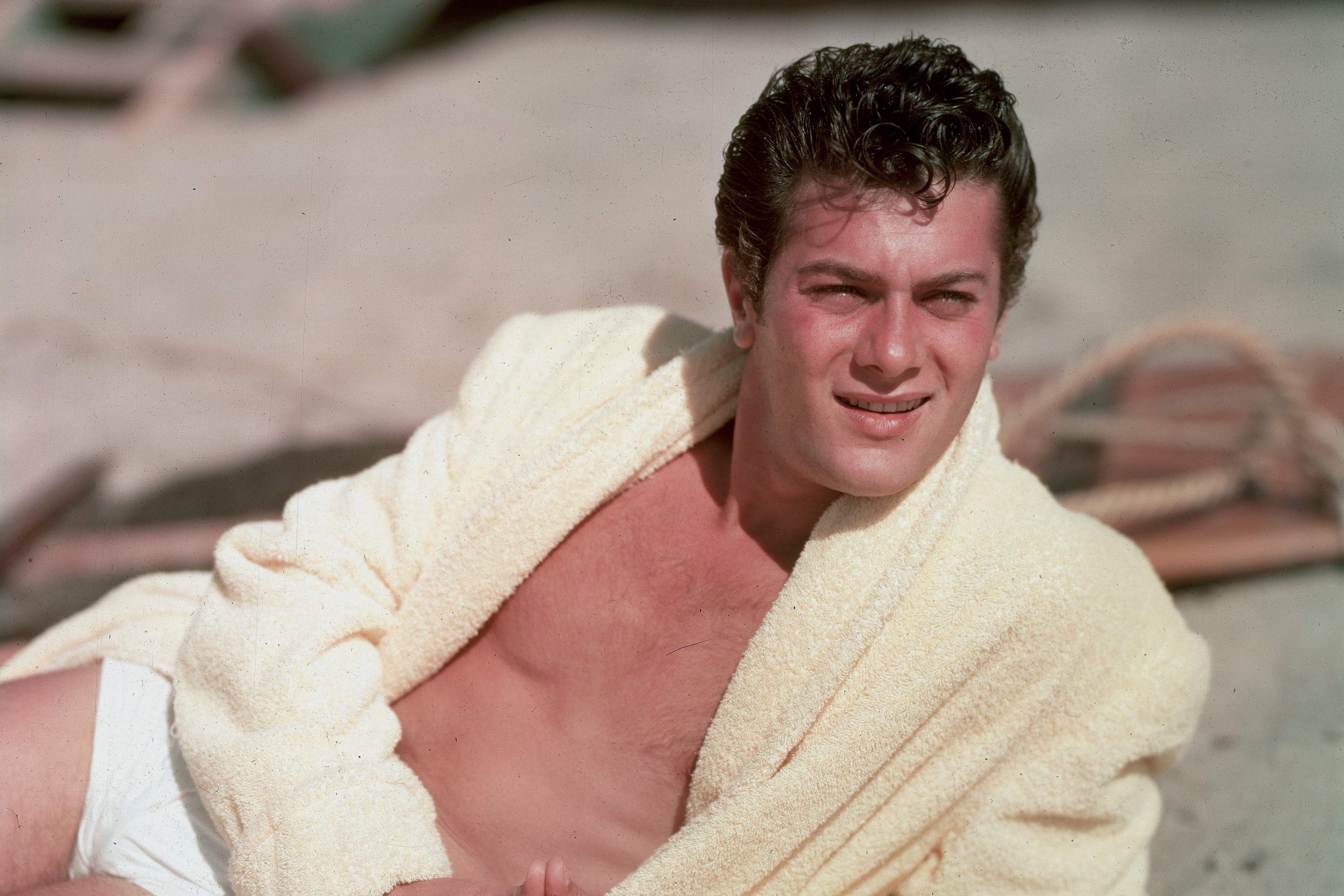 Az amerikai színész, Tony Curtis fürdőköpenyben és fehér úszónadrágban heverészik a tengerparton
