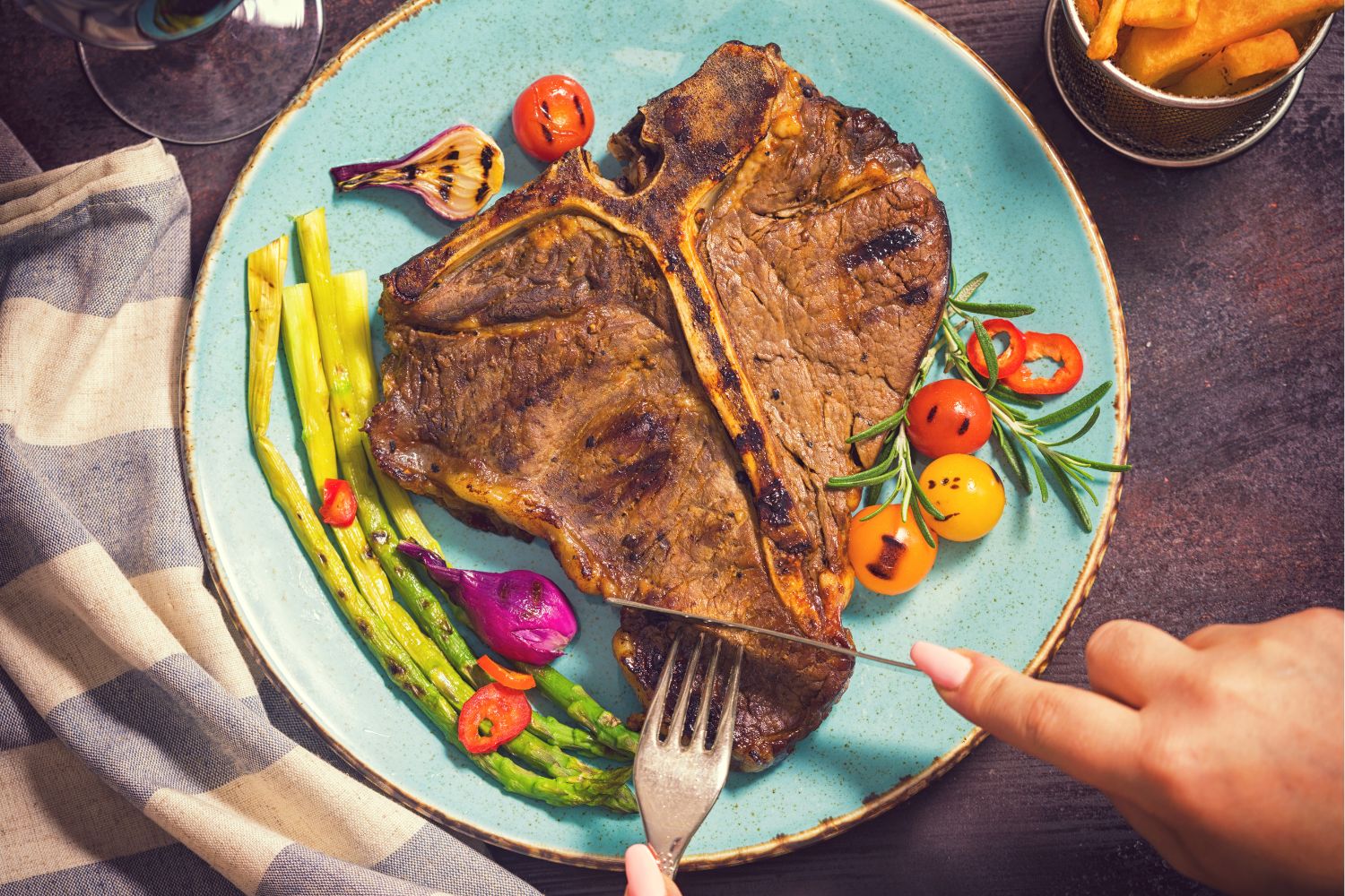 Kék kerámia tányéron színes zöldségek és steak, amit nő eszik késsel és villával