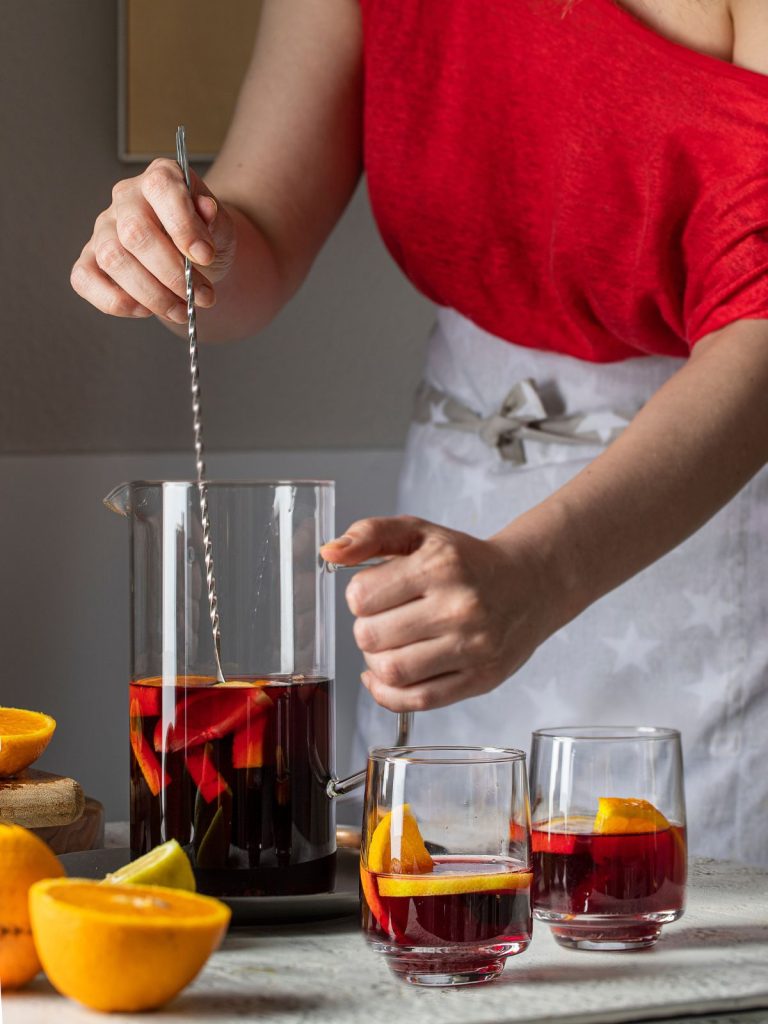 Nő piros felsőben kancsóban tinto de verano spanyol nyári koktélt kever, mellette citromszeletek, üvegpohár a koktéllal