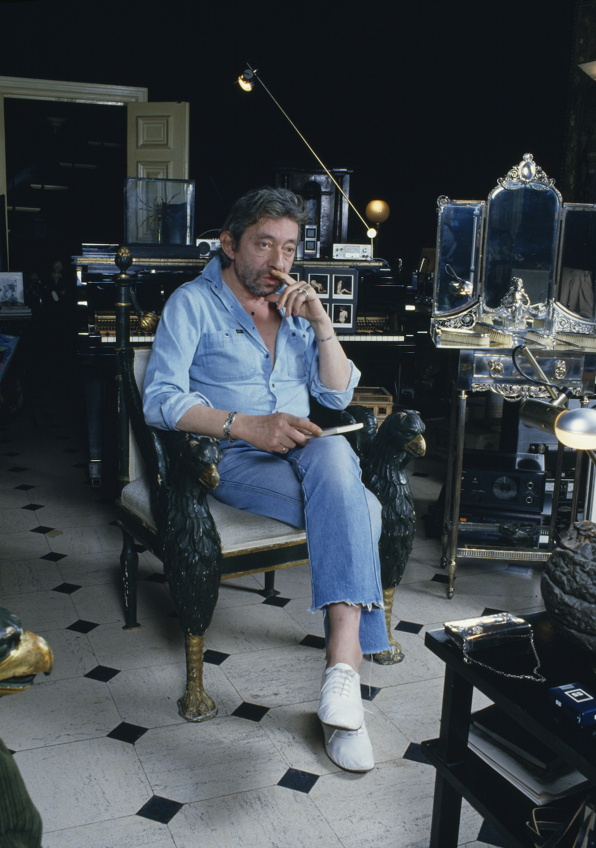 Serge Gainsbourg kék farmeringben és nadrágban, fehér cipőben ül egy kávézóban