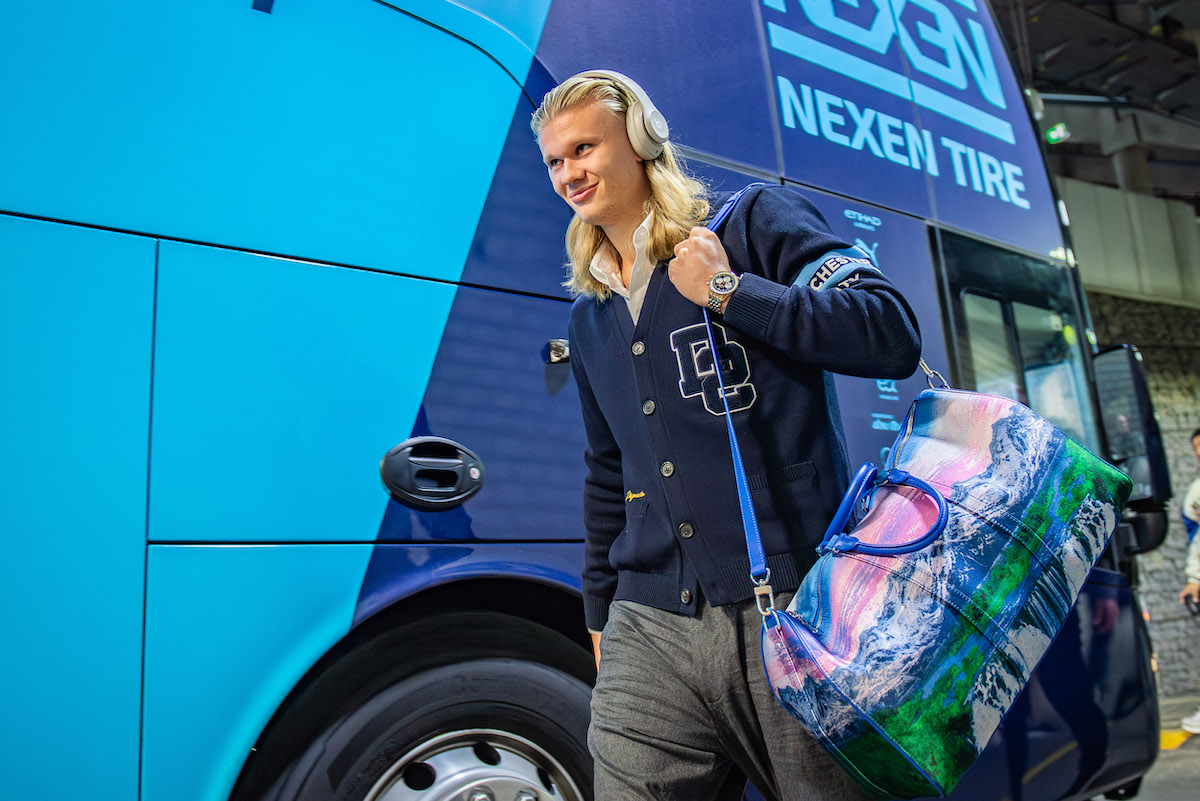 Erling Haaland kék háttér előtt fülesben, színes táskával