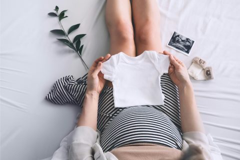 Várandós nő padlón ül csíkos ruhában, ölében fehér rugdalódzó, mellette eukaliptuszág és ultrahangos kép, felülnézetből fotózva