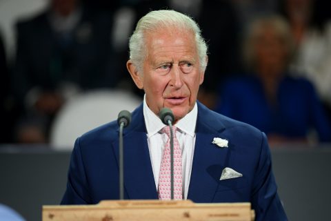 Károly király mikrofonnál beszél, sötétkék öltönyben és rózsaszín nyakkendőben