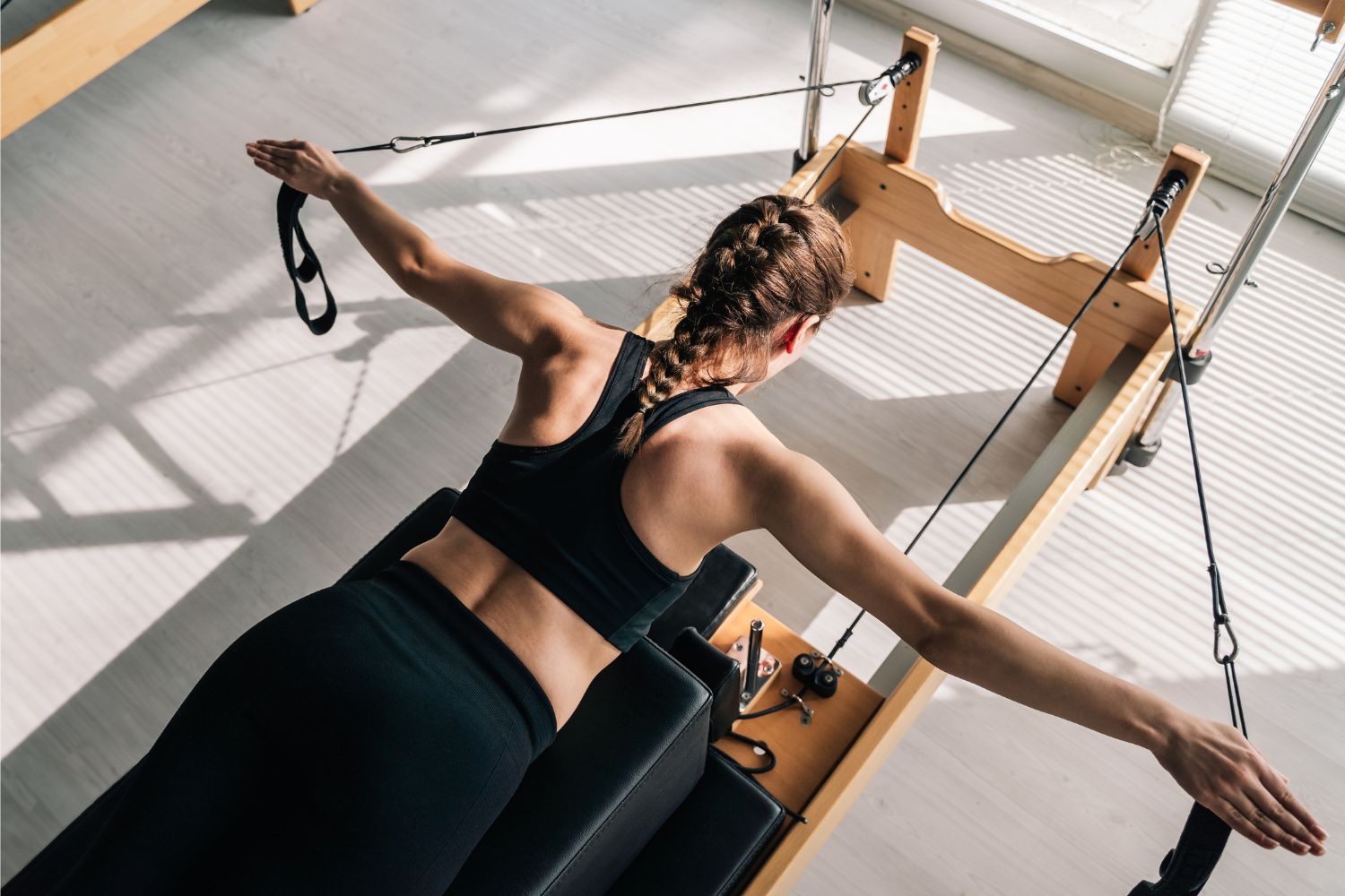 Nő hason fekszik pilates reformer gépen fonott hajjal fekete edzőruhában