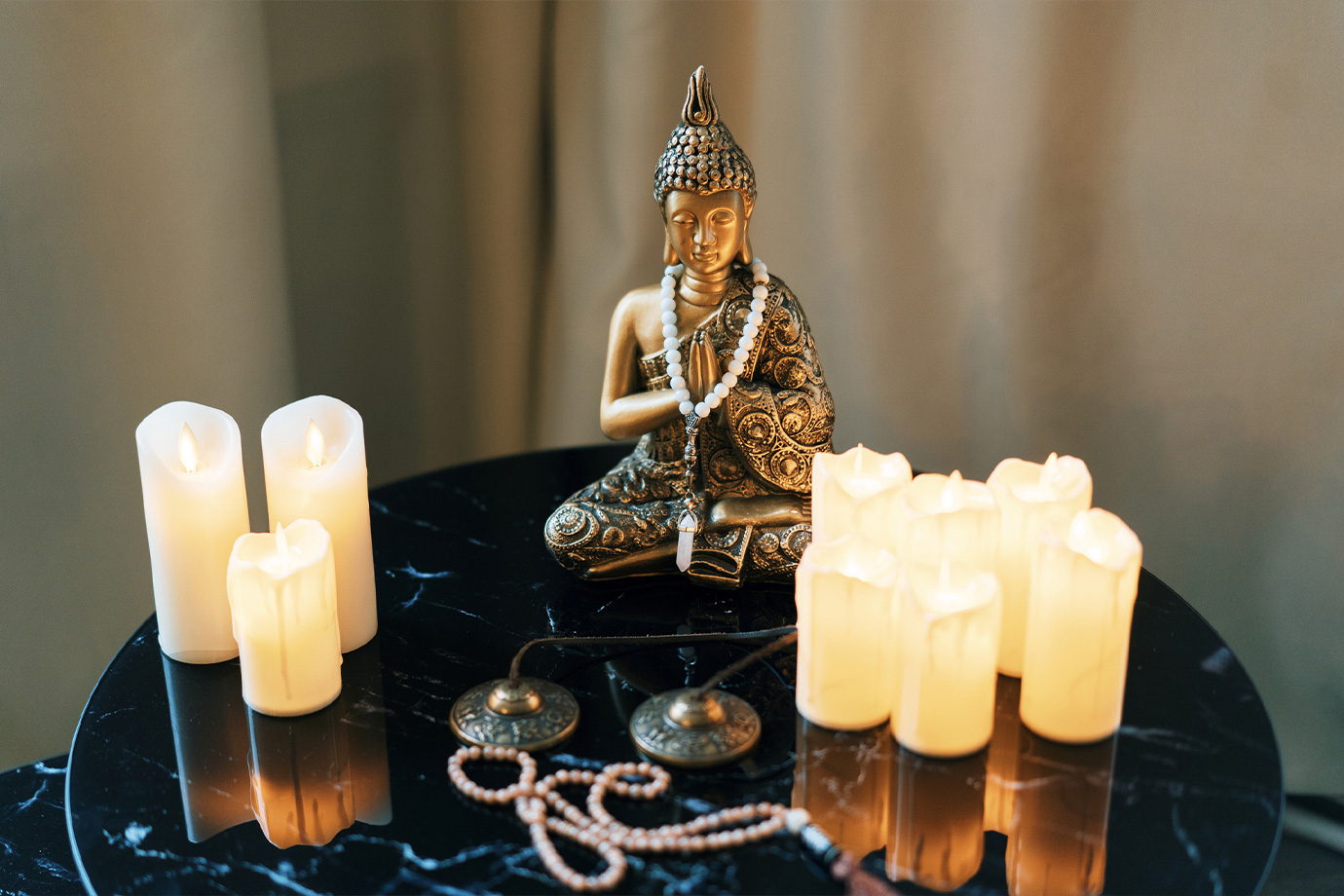 Arany Buddha-szobor egy fekete márványasztalon ül, körülötte égő gyertyák, imafüzérek és tingsha cintányérok