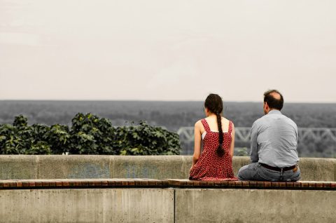 háttal ülő pár, előttük fák és horizont, a nő piros nyári ruhát és fonatot visel