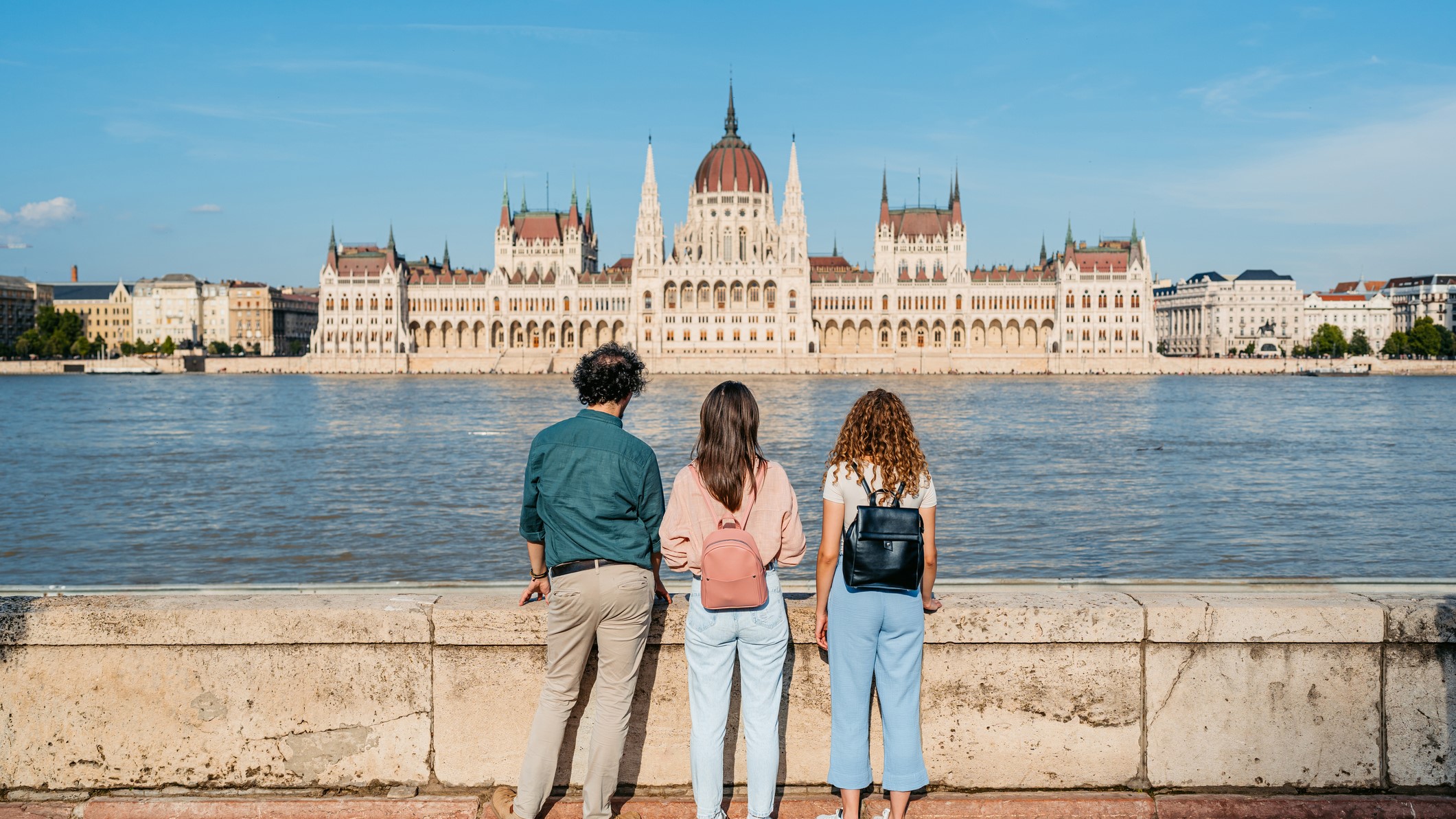 kulfoldon-elo-magyarok-hazateres-kormanyvaltas | Marie Claire Három barátból álló csoport, akik a Duna túloldalán, a magyarországi Budapest városában a magyar parlament épületére néznek.