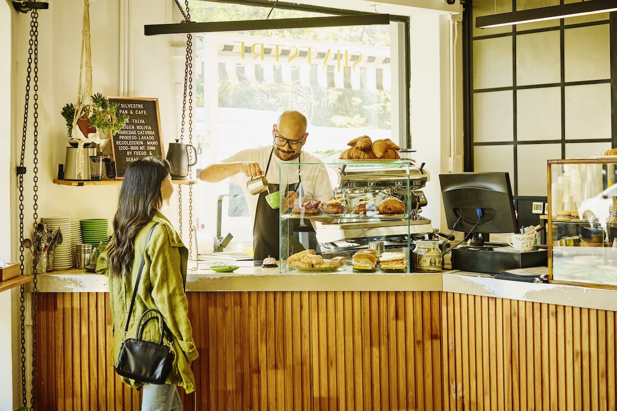 munkahelyi-ebed-etikett | Marie Claire barista kávézó pultjában kávét készít, előtte hosszú hajú nő várakozik