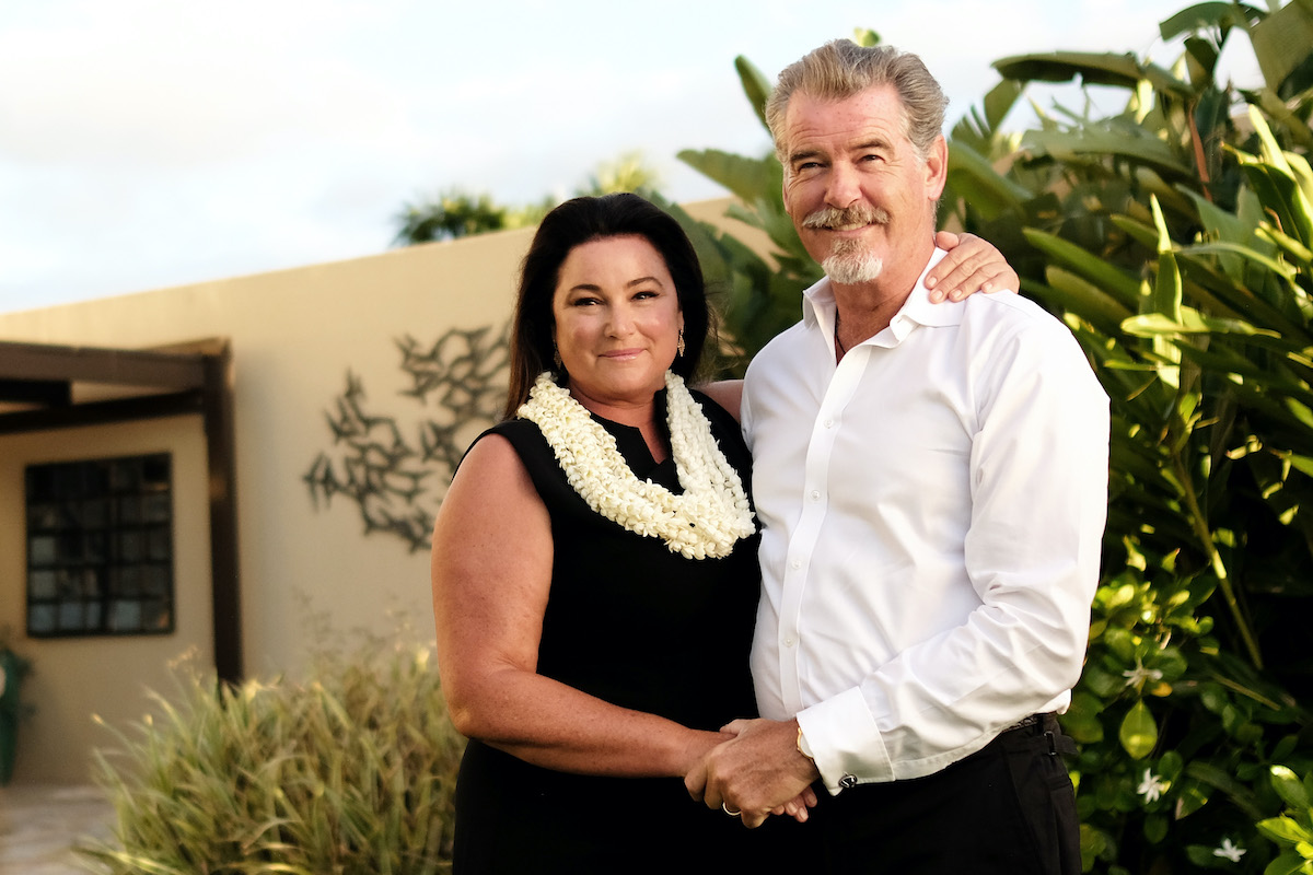 Keely Shaye Brosnan fekete ruhában, nyakában fehér füzér és Pierce Brosnan szakállal, fehér ingben