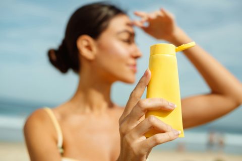 barna hajú nő a strandon, naptejes flakont tart a kamera felé
