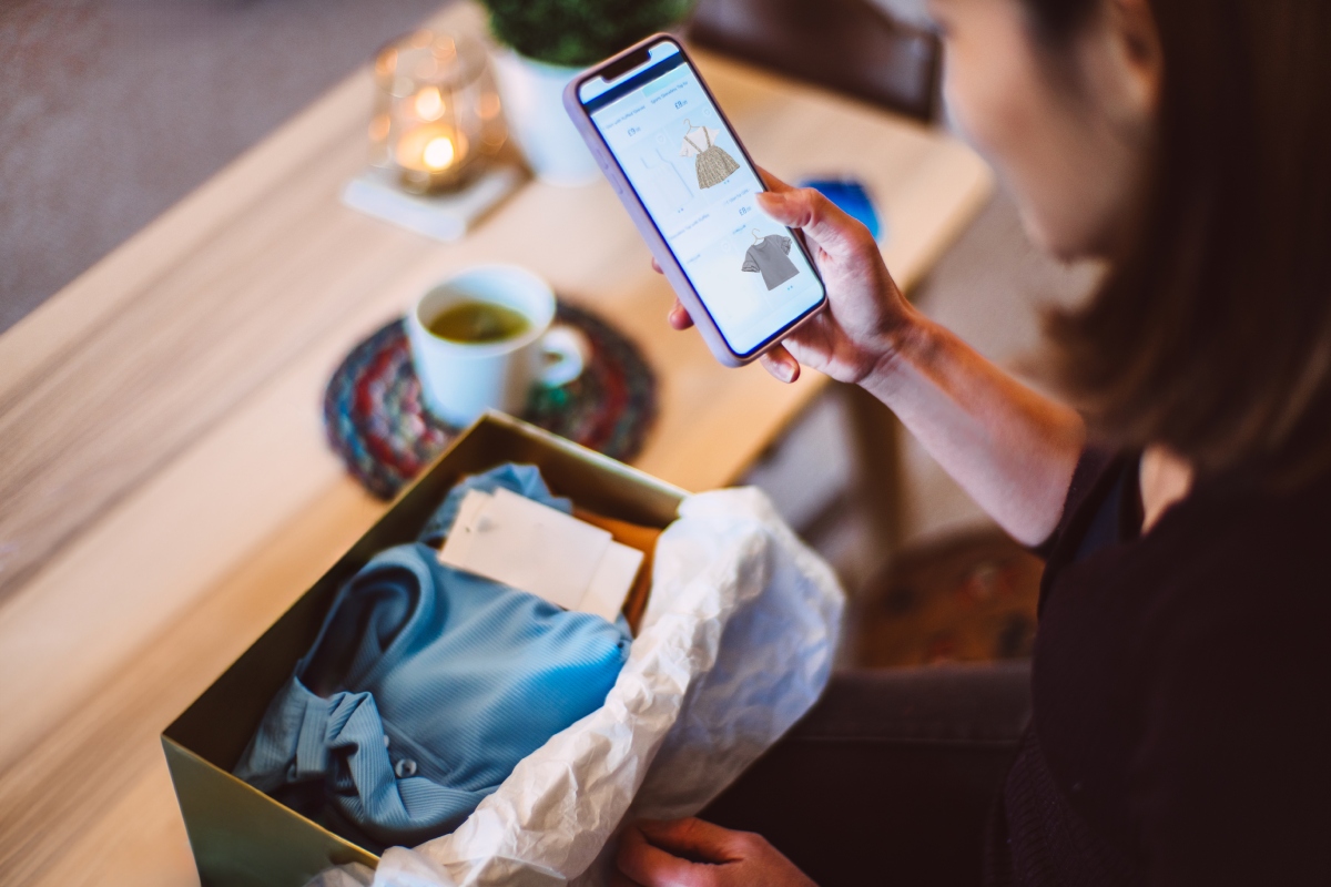 mergezo-ruha-fast-fashion-anyagok | Marie Claire Fiatal nő otthon, okostelefonon vásárol ruhákat online. Az online boltból kiszállított csomag ott van előtte kikészítve.