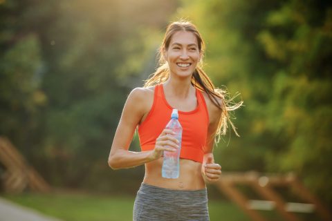 Nő narancsszínű sportmelltartóban és szürke leggingsben gyalogol parkban vizespalackkal a kezében