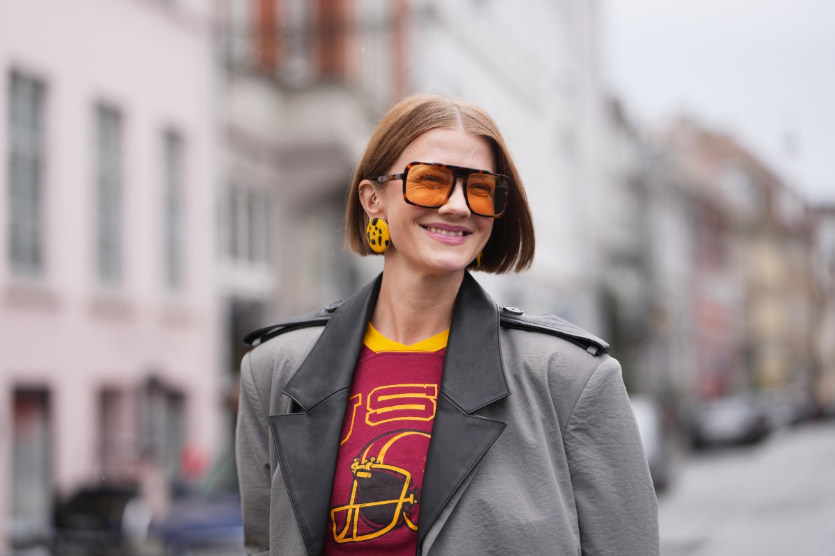 Marianne Theodorsen világosbarna, állig érő, egyenes szálú, középen elválasztott bobfrizurát, oversized szögletes fekete napszemüveget narancsszínű lencsékkel, sárga-fekete fülbevalót, fekete bőrhajtókás, nyitva hagyott szürke ballonkabátot és piros, grafikás, kerek nyakú pólót visel az MKDT show előtt, a 2027-es tavaszi-nyári Copenhagen Fashion Week idején.