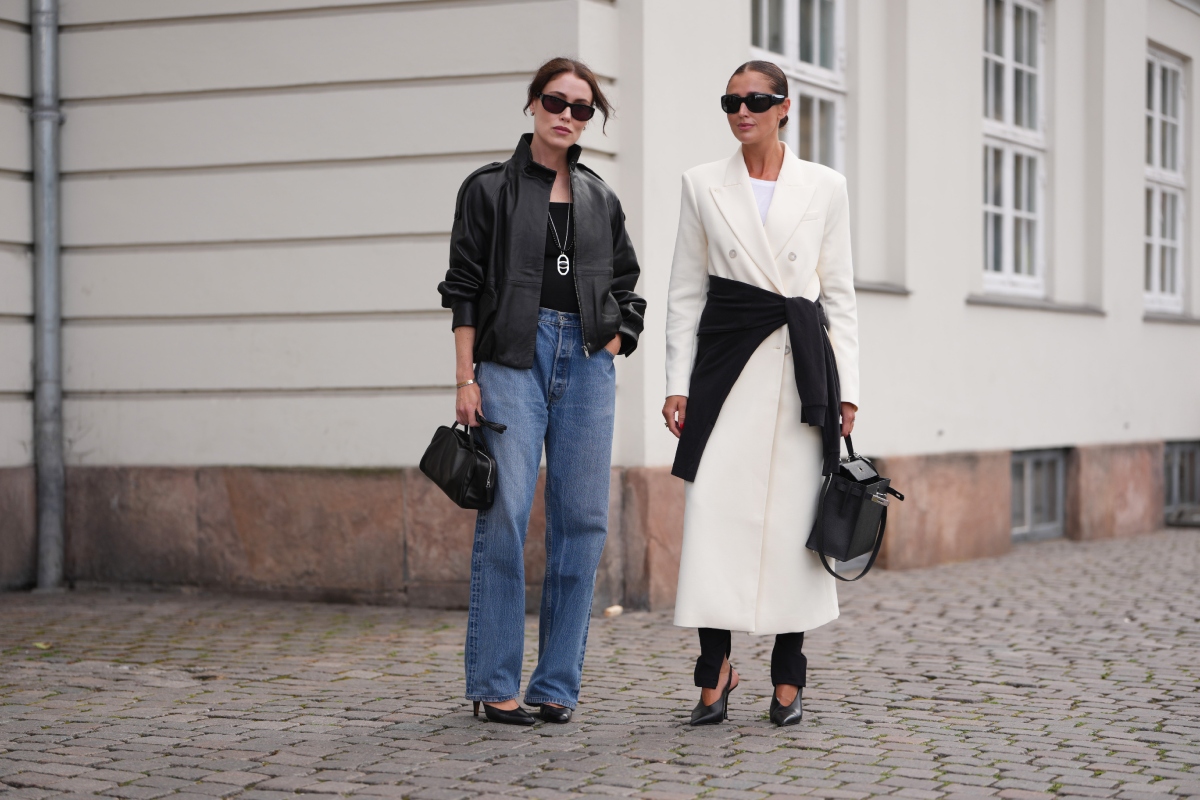 stilusos-nok-kevesebb-ruha-vasarlas-kapszulagardrob | Marie Claire Annabel Rosendahl barna haját alacsony kontyban, fekete szögletes napszemüveget sötét lencsékkel, ezüst nyakláncot, fekete bőr bomberdzsekit, fekete felsőt, kék egyenes szárú farmert, fekete, felül fogantyús táskát és fekete, hegyes orrú cipőt visel; Darja Barannik hátrasimított, középen elválasztott sötétbarna haját, fekete napszemüveget, fehér pólóra rétegzett krémfehér, kétsoros, hosszított blézert, fekete nadrágot és derékon megkötött fekete pulóvert visel az Almada Label show előtt, a 2027-es tavaszi-nyári Copenhagen Fashion Week idején, 2026. augusztus 5-én, Koppenhágában.