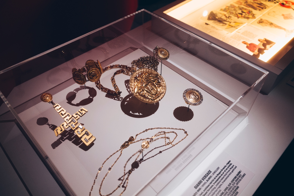 versace-retrospektiv-kiallitas-parizs-2026 | Marie Claire Vitrinben aranyszínű Versace-ékszerek: Medúza-fejes medál, meanderes kereszt és láncok.