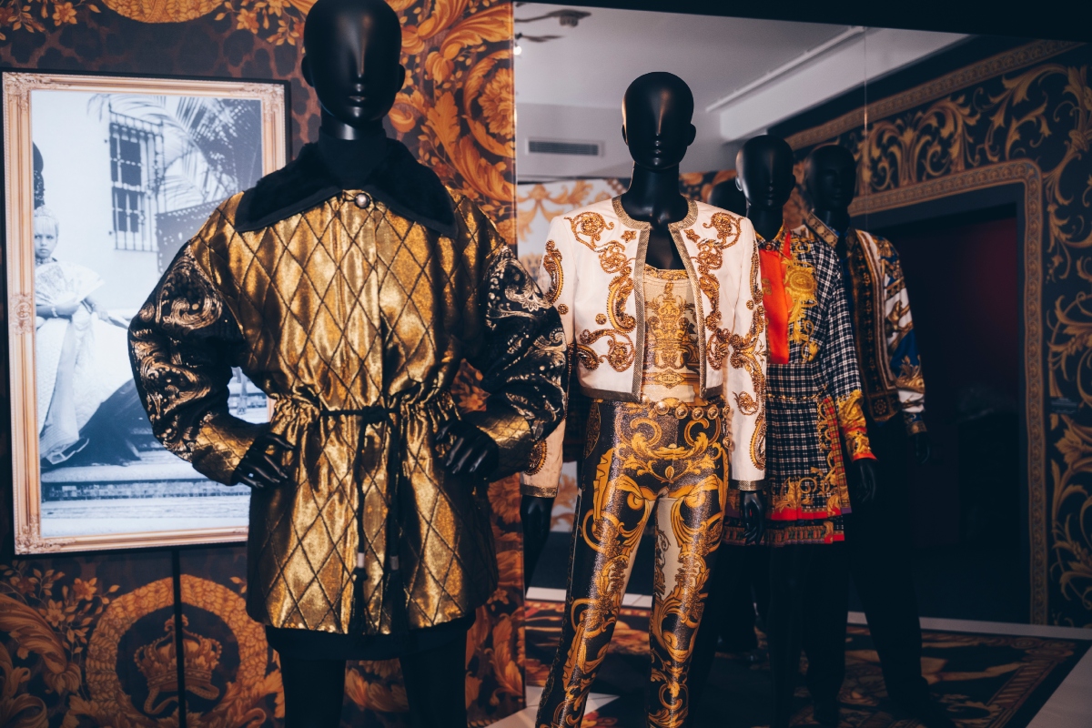 versace-retrospektiv-kiallitas-parizs-2026 | Marie Claire Aranyszínű steppelt kabát bábun, mögötte barokk mintás Versace-ruhák sora a kiállításon.