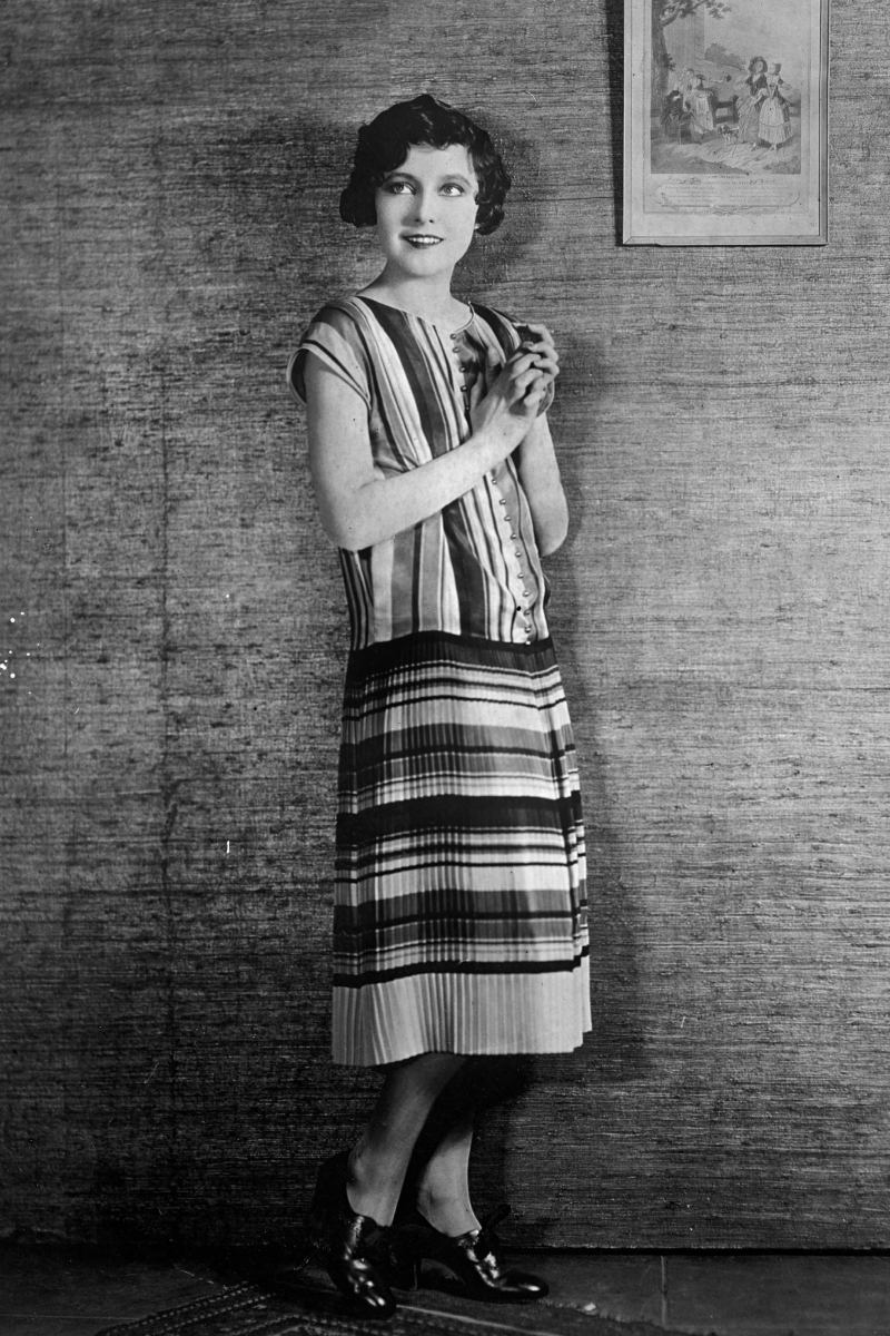 Betty Bronson amerikai színésznő 1925 körül, narancs, zöld, piros és rézszínű, római csíkos, rakott kötött szoknyában, sárgaréz gombokkal.