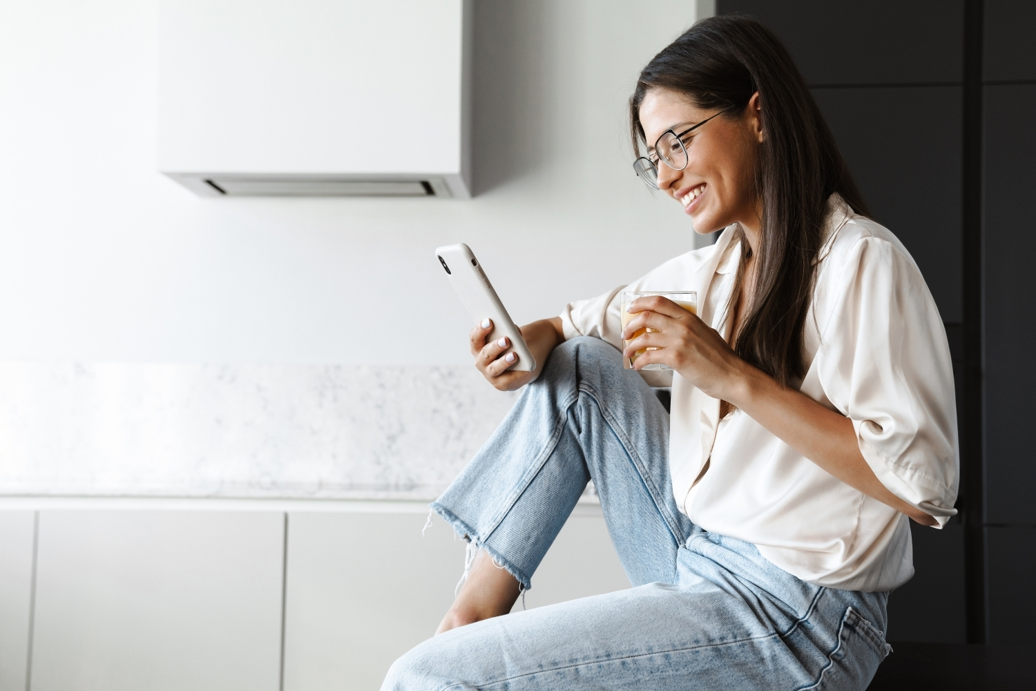 Nézzük meg újra | Marie Claire Nő farmerben és kényelmes felsőben mosolyogva nézi a telefonját