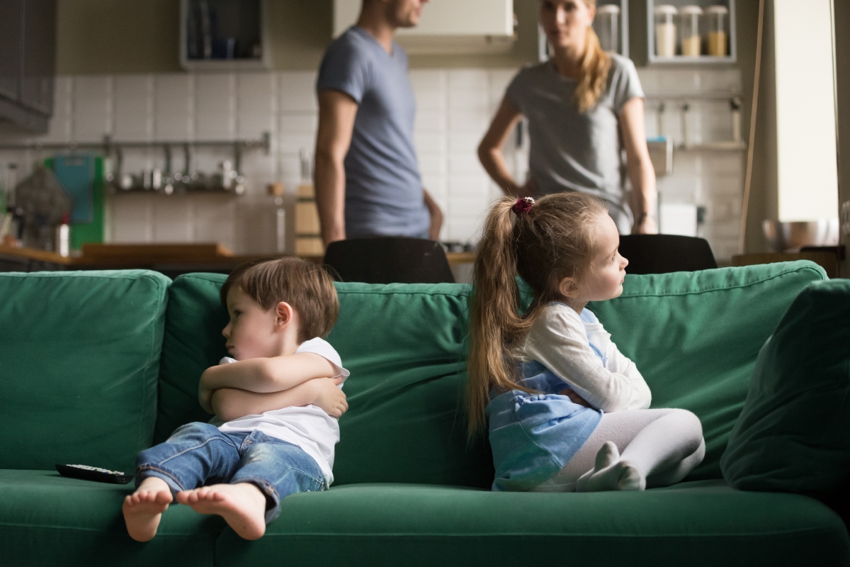 kettos-merce-lanyok-fiuk-anya-vallomas | Marie Claire Zöld kanapén ül egy kisfiú és egy kislány, nem néznek egymásra, a háttérben a szüleik