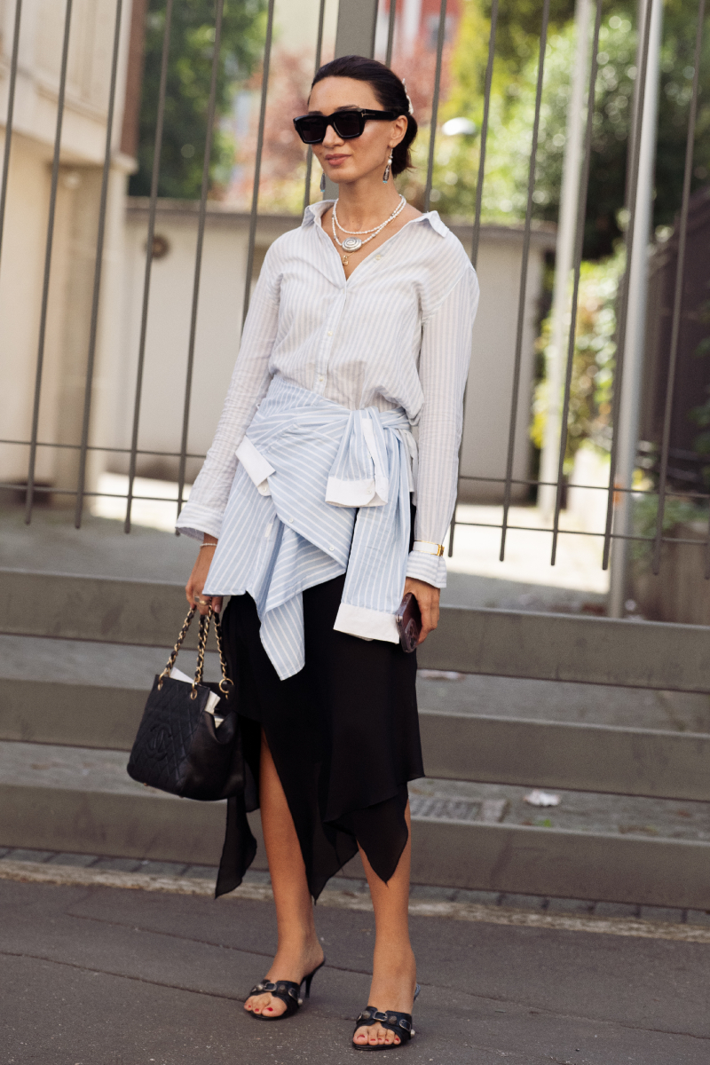 milanoi-divathet-2026-streetstyle-midiszoknya | Marie Claire Vendég halványkék csíkos ingben, derekán ugyanolyan inggel megkötve, fekete, aszimmetrikus szatén midiszoknyában, fekete magas sarkú szandálban, steppelt fekete táskával a Dhruv Kapoor bemutatója előtt a milánói divathéten.