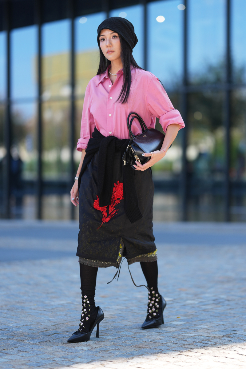 milanoi-divathet-2026-streetstyle-midiszoknya | Marie Claire Yoyo Cao rózsaszín, felhajtott ujjú ingben, piros hímzéses fekete midiszoknyában, derekára kötött fekete pulóverrel, fekete kötött sapkában, virágmintás harisnyában és fekete Prada táskával a Prada bemutatója előtt a milánói divathéten.