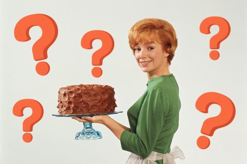 Retró fotó vöröshajú nő zöld felsőben, fehér kötényben, tortaállványon csokoládé tortát tart