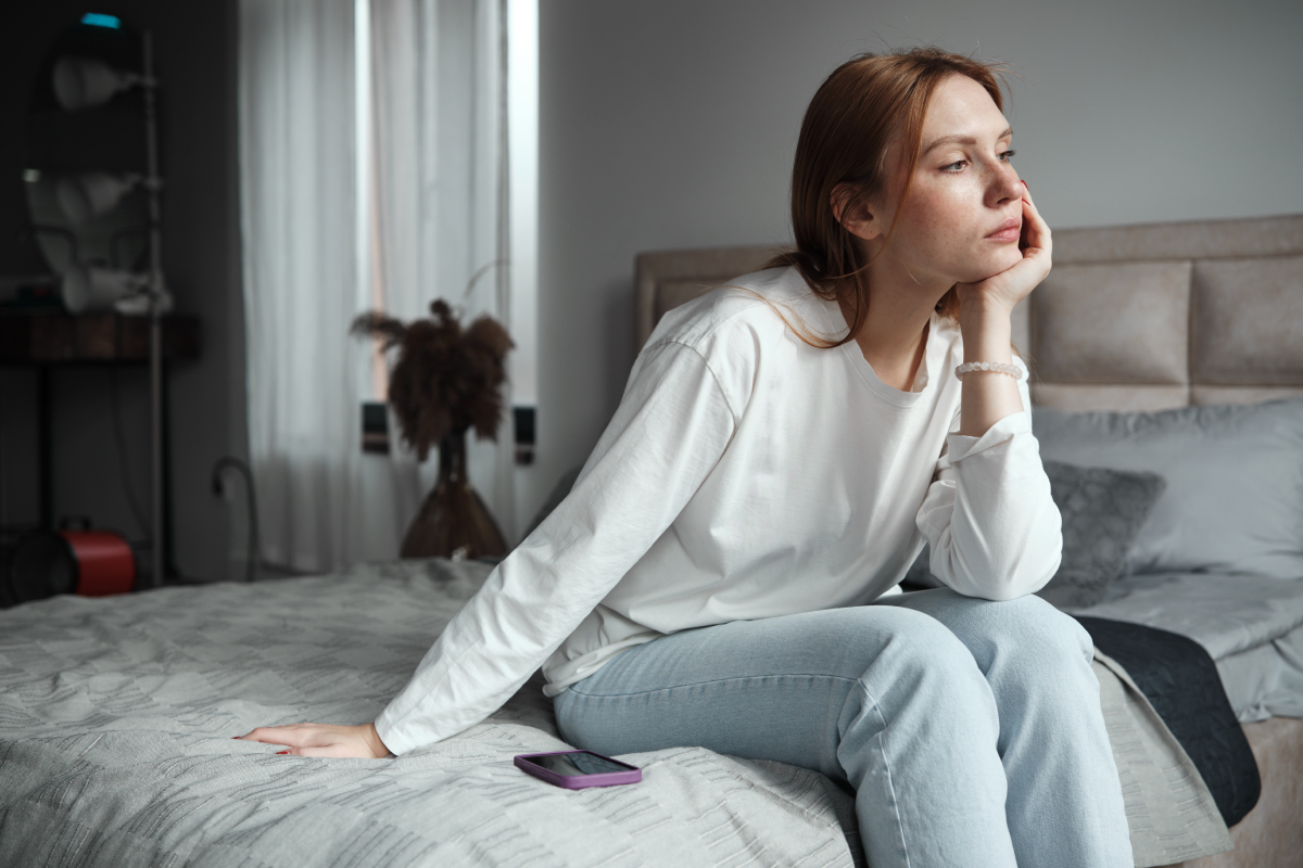situationship-hogyan-lepj-tul-rajta-ha-sosem-voltatok-egyutt | Marie Claire Egy fiatal nő farmerben, pólóban ül az ágy szélén szomorúan, mellette telefon
