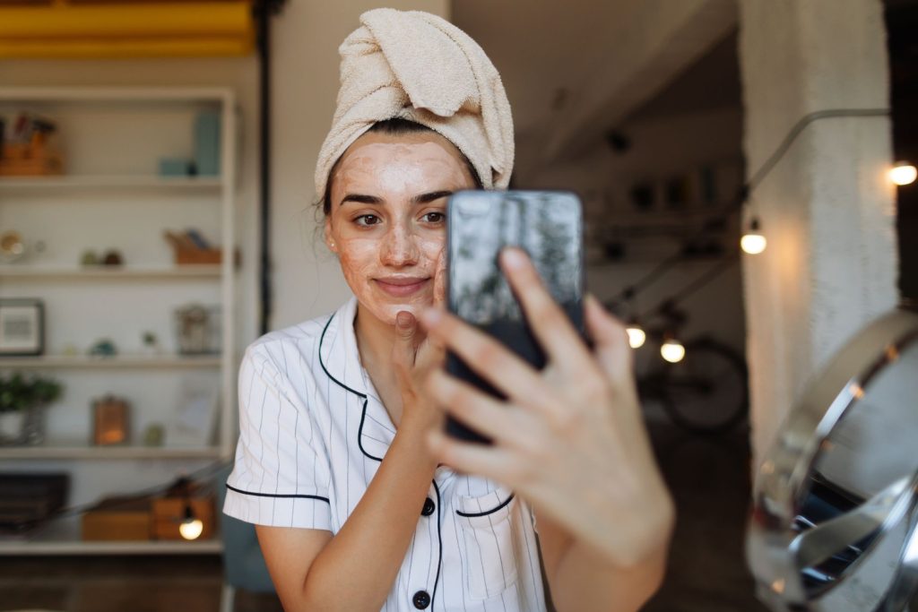 Fiatal nő krémezi az arcát, közben telefonon nézi magát vagy szelfizik - skintellectual trend