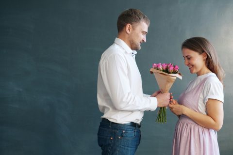rózsaszín ruhás nőnek férfi rózsaszín csokrot nyújt át szürke háttér előtt