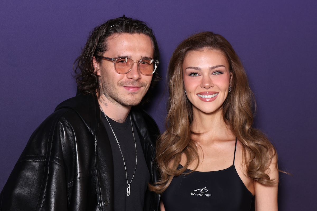 Brooklyn Beckham szemüvegben és fekete bőrdzsekiben és Nicola Peltz fekete topban, lila háttér előtt