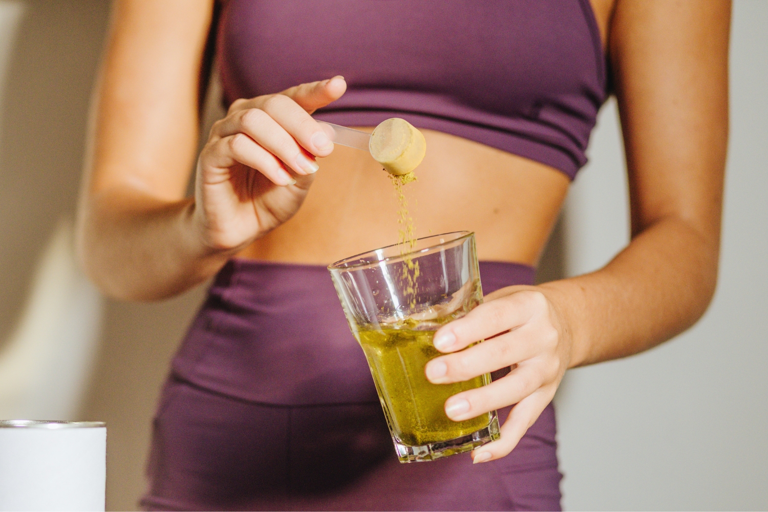 Nő lila fitneszszettben étrend-kiegészítő port önt mérőkanálból egy pohár vízbe